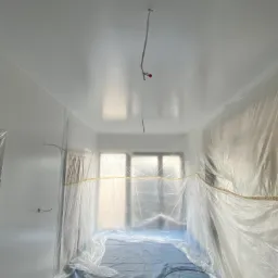 Pokój przygotowany do prac wykończeniowych, zabezpieczony folią malarską i taśmą, świeżo pomalowane ściany i sufit na biało, widoczne wystające przewody elektryczne.