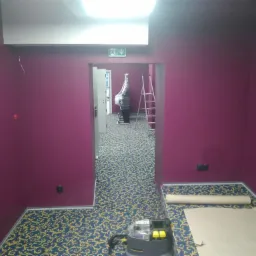 Wnętrze pomalowane na fioletowo z wzorzystą wykładziną, widoczny odkurzacz piorący, zabezpieczona podłoga i drabina w przejściu do kolejnego pomieszczenia.