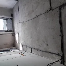 Surowe wnętrze w trakcie remontu z widocznymi przewodami elektrycznymi przymocowanymi do betonowej ściany za pomocą metalowych uchwytów. Podłoga wyłożona białymi płytami izolacyjnymi, a w tle okno...