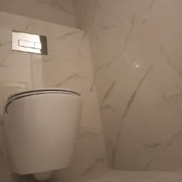 Podwieszana toaleta z chromowanym przyciskiem spłukującym na ścianie wyłożonej białymi płytkami imitującymi marmur.