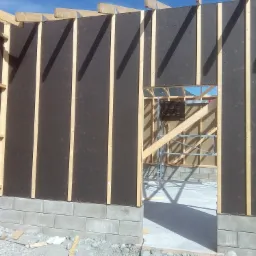 Konstrukcja ściany zewnętrznej budynku w trakcie budowy, widoczne płyty poszycia i drewniane słupki, fundament z bloczków betonowych, w tle fragment konstrukcji drewnianej i niebieskie niebo.