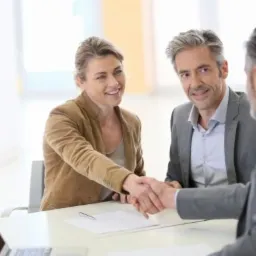 Uśmiechnięta kobieta w brązowej marynarce i mężczyzna w szarym garniturze uściskiem dłoni finalizują umowę kredytową przy biurku z laptopem i dokumentami.