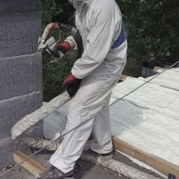 Pracownik w kombinezonie i masce ochronnej aplikuje izolację na dachu, widoczny fragment komina z szarych bloczków i świeżo nałożona, jasna pianka izolacyjna.