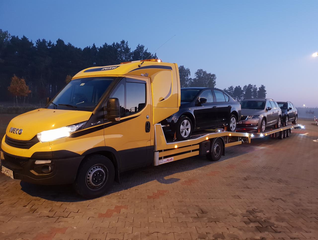 Żółta laweta Iveco z trzema samochodami marki Skoda na platformie, transportowana w Gdańsku o zmierzchu.