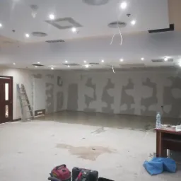 Wnętrze w trakcie prac przygotowawczych do remontu: ściany z widocznymi śladami szpachlowania, surowe podłoże, drabina oparta o ścianę, widoczne narzędzia i materiały budowlane, wiszące kable...