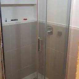 Kabina prysznicowa z przesuwnymi drzwiami w łazience wykończonej szarymi płytkami, widoczna półka z akcesoriami i odpływ liniowy w podłodze.