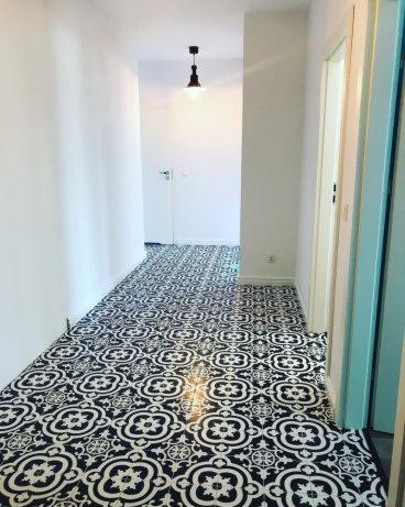 Korytarz z nowo ułożoną, wzorzystą, czarno-białą podłogą z płytek ceramicznych, widok w perspektywie, z wiszącą czarną lampą w tle i białymi ścianami.