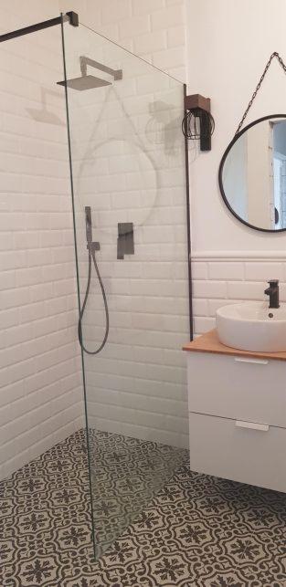 Nowoczesna łazienka z kabiną prysznicową typu walk-in, białe płytki imitujące cegłę, wzorzysta podłoga, okrągłe lustro na łańcuszku i minimalistyczna umywalka nablatowa.