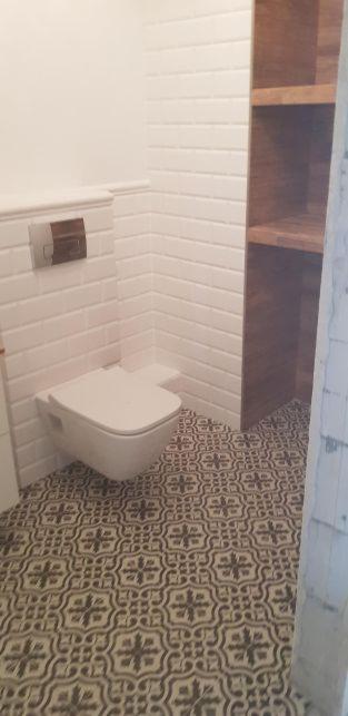 Nowoczesna toaleta wisząca z podtynkowym systemem spłukiwania, białe kafelki typu metro na ścianach, wzorzysta podłoga i drewniana półka w łazience.