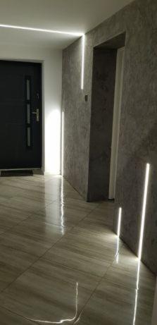 Korytarz z szarymi ścianami o nieregularnej fakturze, podświetlony liniowym oświetleniem LED wzdłuż ścian i sufitu, z błyszczącą, beżową podłogą imitującą drewno i czarnymi drzwiami wejściowymi.