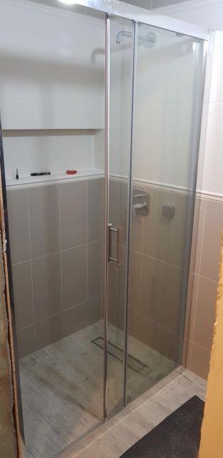 Kabina prysznicowa z przesuwnymi drzwiami w łazience wykończonej szarymi płytkami, widoczna półka z akcesoriami i odpływ liniowy w podłodze.