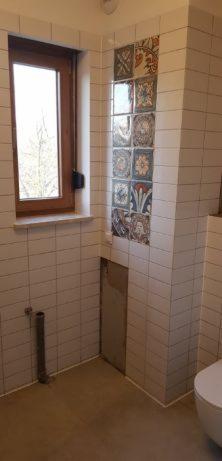 Wnętrze łazienki w trakcie remontu, widoczne białe płytki na ścianach, dekoracyjne płytki nad zabudową, okno z drewnianą ramą oraz wystająca rura kanalizacyjna.