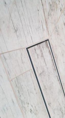 Ułożone jasne panele podłogowe z widocznymi fugami i odpływem liniowym, imitujące strukturę drewna z wyraźnymi słojami.