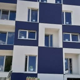 PAXON LTD - Nowoczesna elewacja budynku w szachownicę, z białymi i granatowymi panelami. Widoczne okna z odbiciami chmur. Częściowo widoczne rusztowania u dołu.