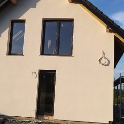 PAXON LTD - Jasna elewacja domu jednorodzinnego z czarnymi oknami i drzwiami. Na ścianie zwisają przewody. Widoczne rusztowania i fragment osoby w prawym dolnym rogu.