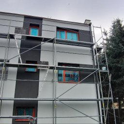 PAXON LTD - Dwukolorowa elewacja domu w trakcie prac. Okna zabezpieczone folią, widoczne rusztowania. Częściowo pomalowana fasada z szarym pasem.