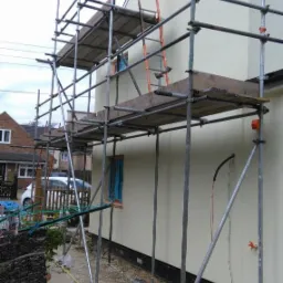 Elewacja domu jednorodzinnego w trakcie prac wykończeniowych, widoczne rusztowanie, drabina oparta o ścianę, świeżo otynkowana ściana w jasnym odcieniu, okno z niebieską ramą i fragment sąsiedniego...