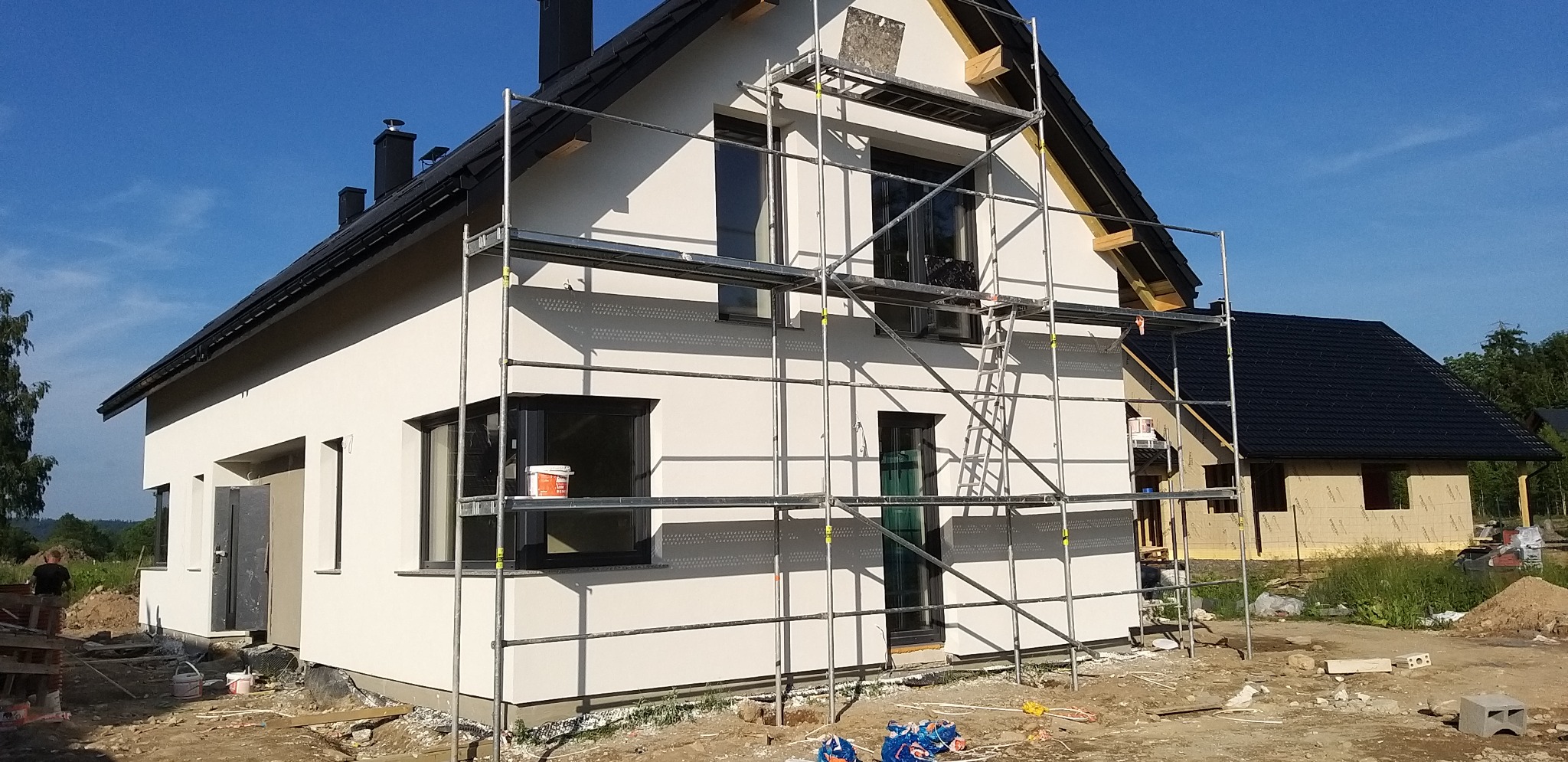 Dom jednorodzinny w trakcie prac elewacyjnych, widoczne rusztowania i wiaderko z farbą na parapecie. Świeżo otynkowana, jasna elewacja. W tle drugi budynek.