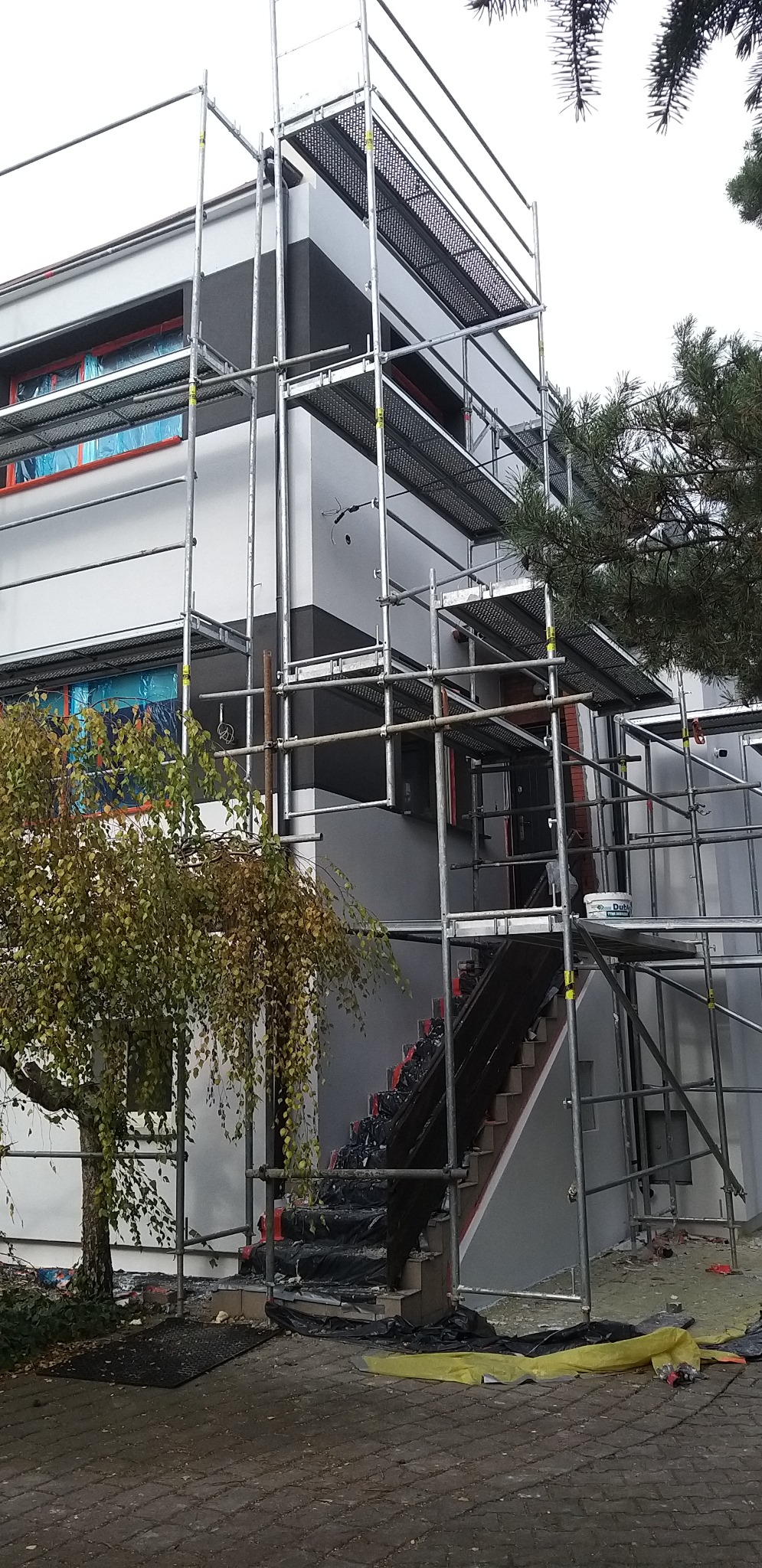 Dwukondygnacyjny budynek w trakcie malowania elewacji, z widocznymi rusztowaniami, zabezpieczonymi oknami i schodami. Częściowo pomalowana ściana na szaro.