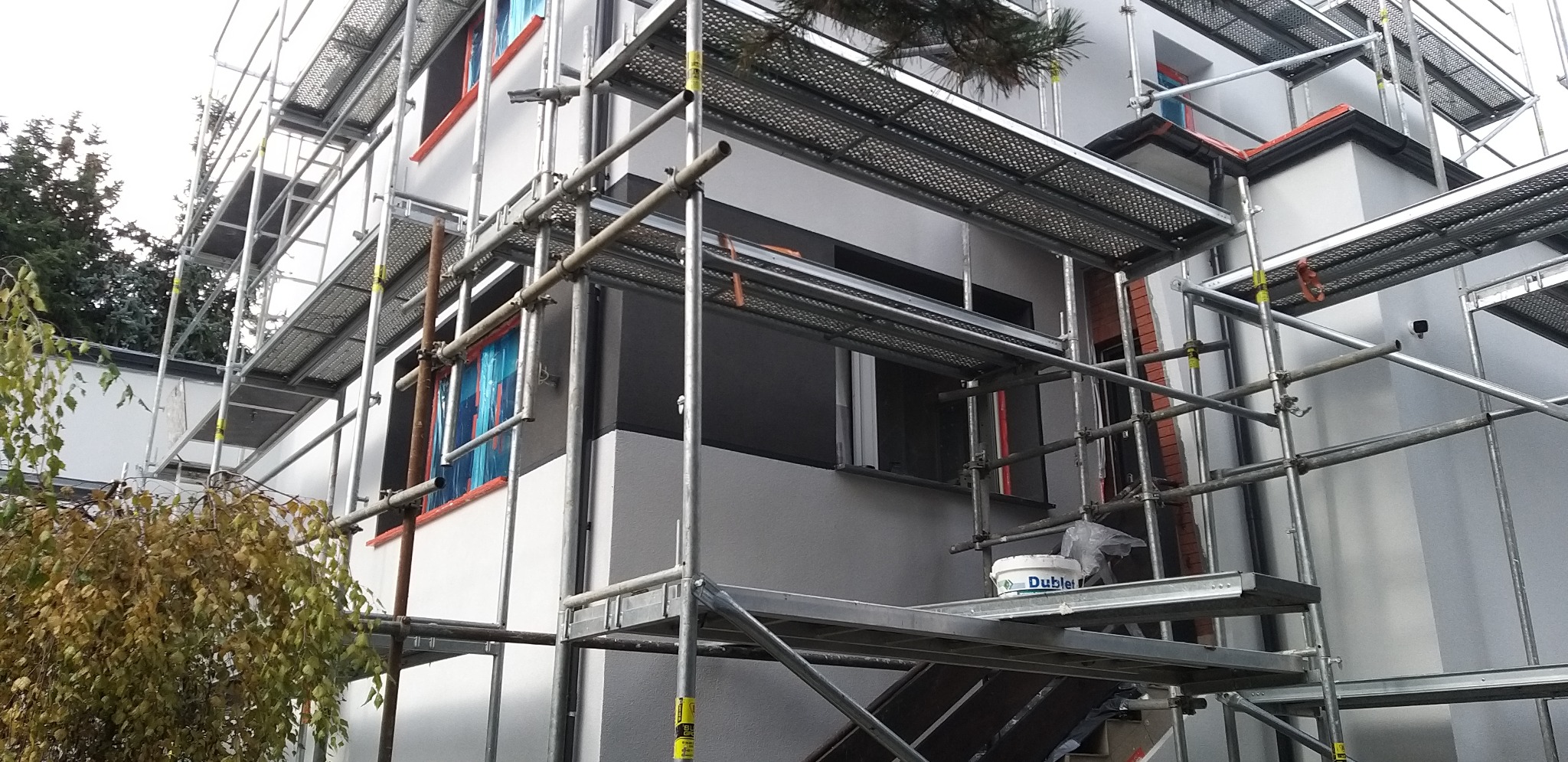 Dom w trakcie malowania elewacji, otoczony rusztowaniem. Widoczne zabezpieczone okna i wiadro z farbą Dublet. Częściowo pomalowana ściana na szaro.