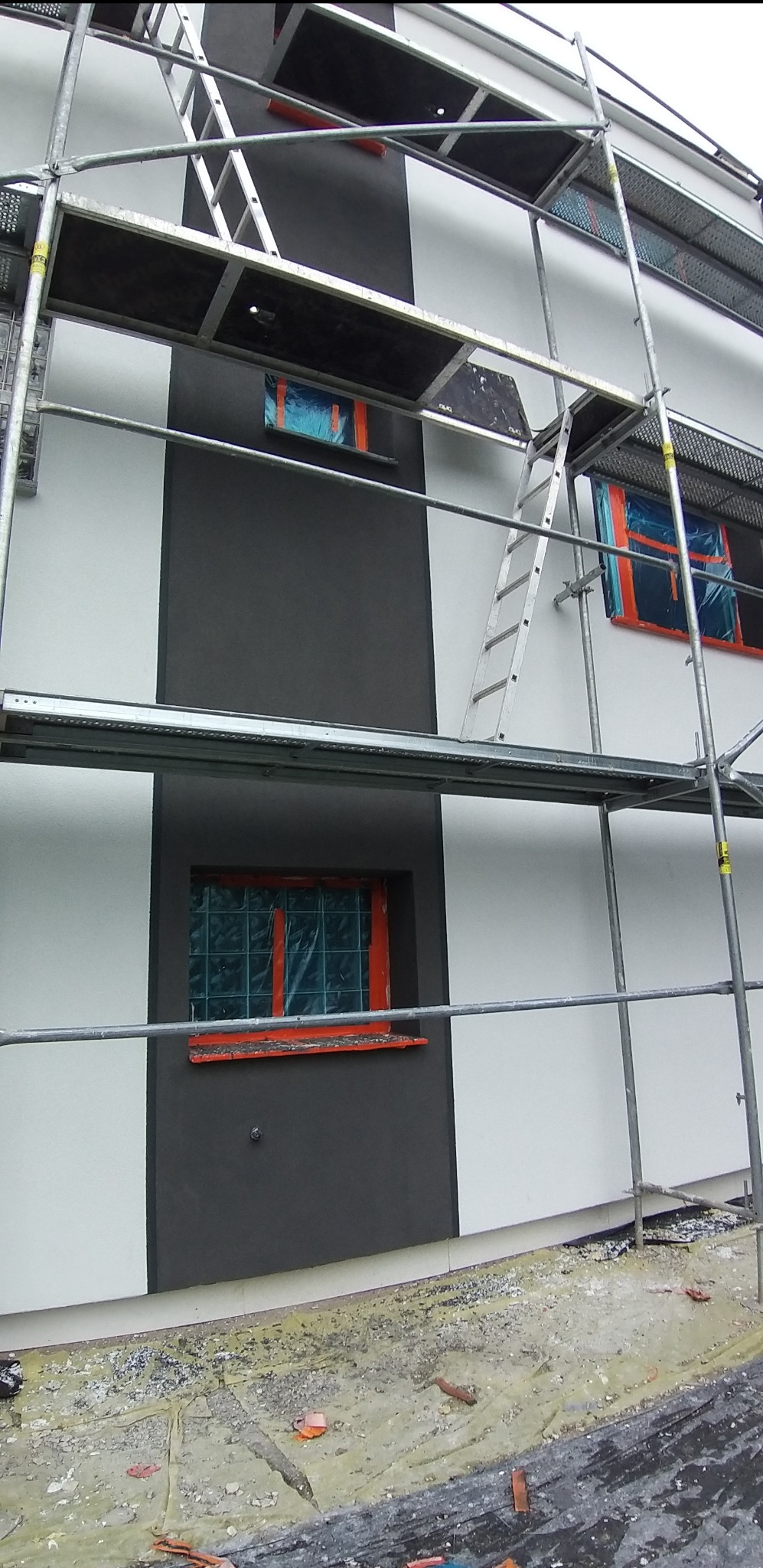 Elewacja budynku w trakcie prac wykończeniowych. Widoczne rusztowania, okna zabezpieczone folią i fragmenty pomalowanej ściany w dwóch kolorach: białym i grafitowym.