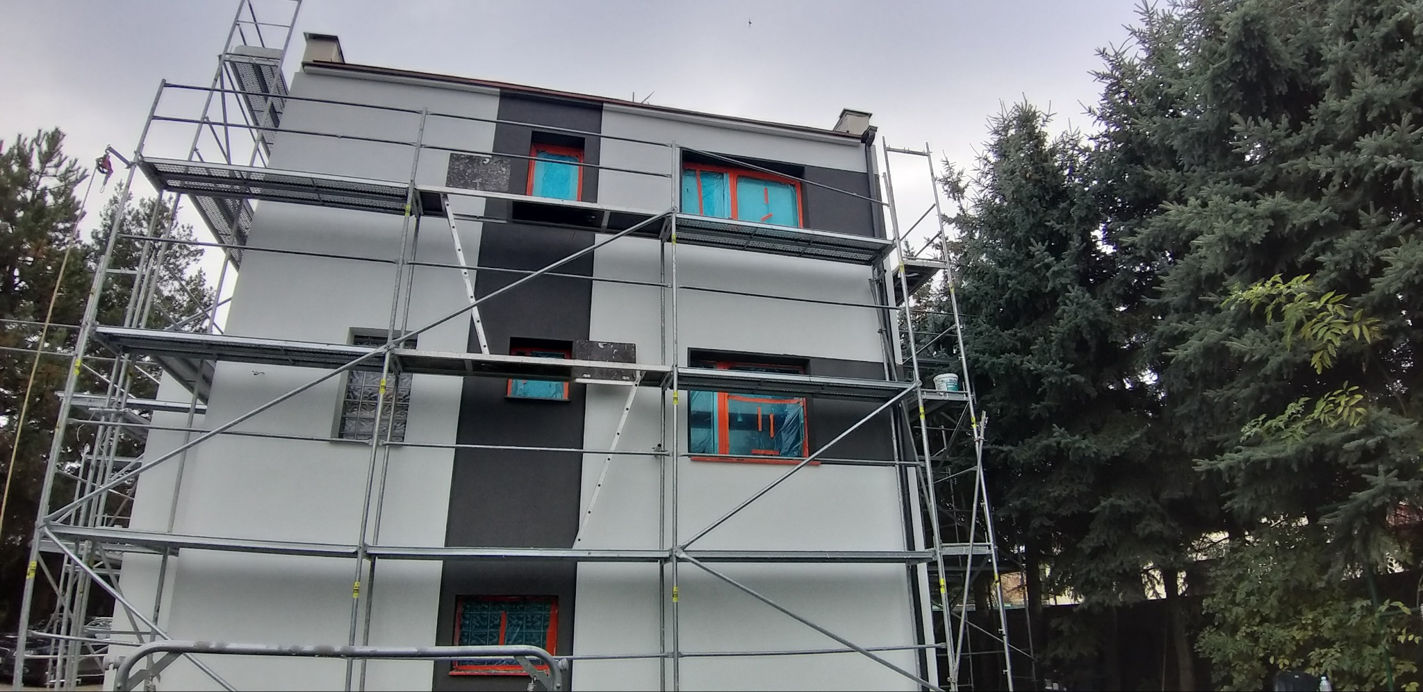 Dwukolorowa elewacja domu w trakcie prac. Okna zabezpieczone folią, widoczne rusztowania. Częściowo pomalowana fasada z szarym pasem.