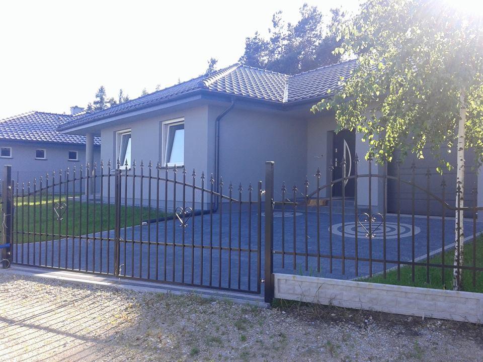 Nowoczesny dom jednorodzinny z szarą elewacją i ciemną dachówką, widoczny zza metalowej bramy z dekoracyjnymi sercami. Podjazd wyłożony kostką brukową z wzorem. Na pierwszym planie fragment...