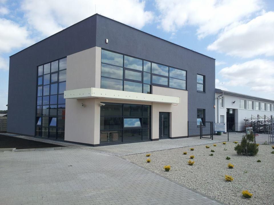 Nowoczesny budynek biurowy z dużymi oknami, pomalowany w odcieniach szarości i beżu, z widocznym wejściem i otoczeniem zagospodarowanym zielenią.