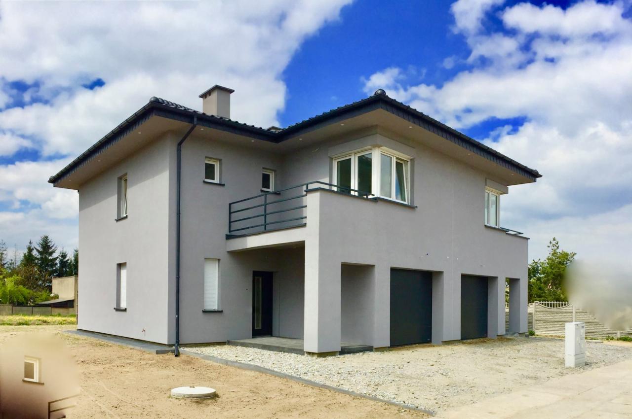 Dwukondygnacyjny dom murowany w odcieniach szarości z balkonem, trzema garażami i ciemnymi bramami garażowymi, na tle błękitnego nieba z chmurami.