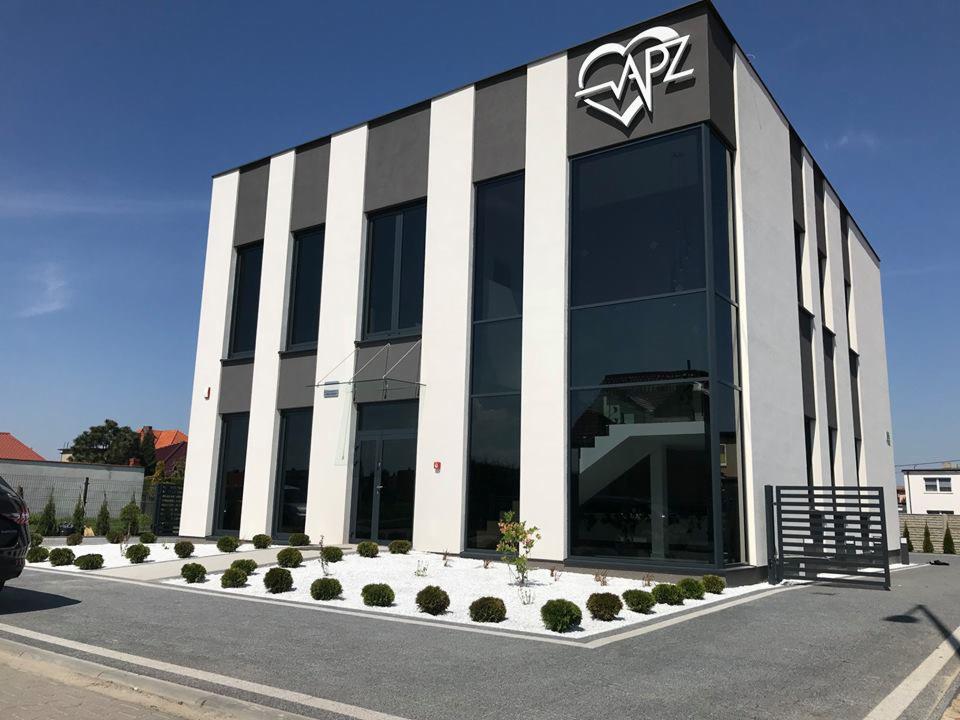 Nowoczesny budynek biurowy z biało-szarymi pasami na elewacji, dużymi oknami i logo APZ na dachu, otoczony białym żwirem i niskimi krzewami.