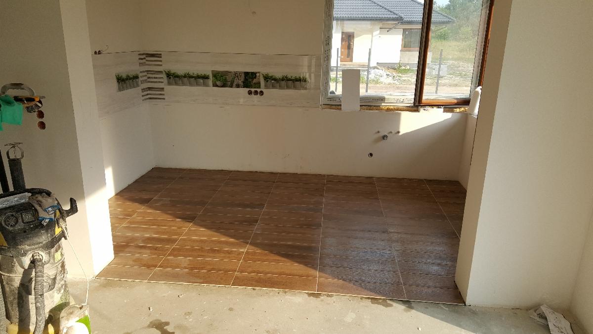 Wnętrze w trakcie remontu: ułożona brązowa podłoga z płytek, częściowo wykończona ściana z dekoracyjnym pasem płytek z motywem roślinnym, widoczne przyłącza wodne i odkurzacz przemysłowy.