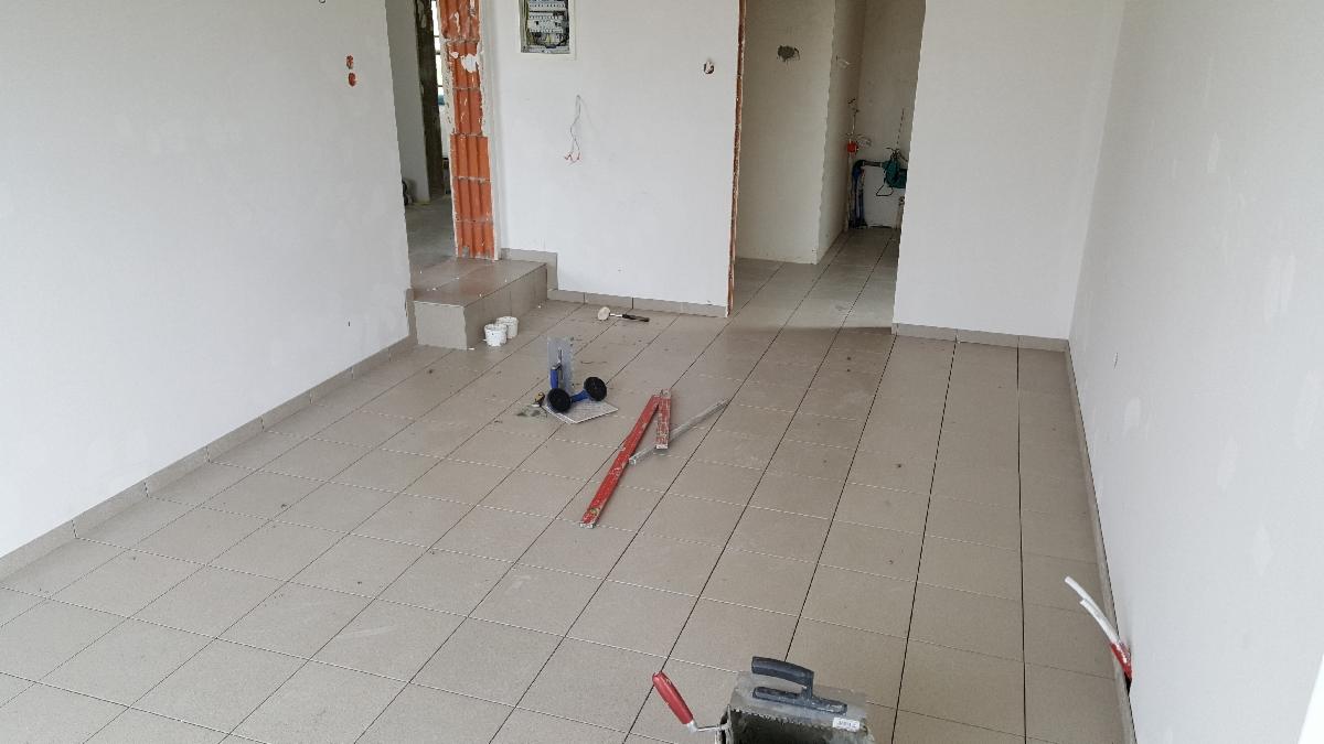 Wnętrze w trakcie układania jasnych, kwadratowych płytek podłogowych; widoczne narzędzia glazurnicze: poziomica, wózek do fugowania, kielnia, oraz niedokończone przejście do innego pomieszczenia...