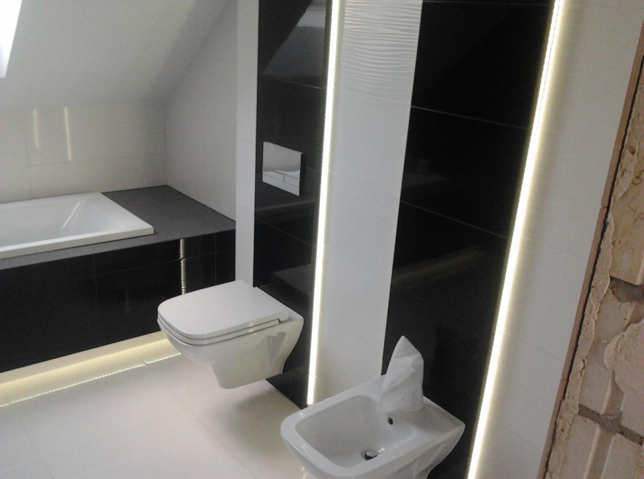Nowoczesna łazienka z czarno-białymi płytkami, podświetleniem LED pod wanną i przy ścianach, wisząca toaleta i bidet, widoczny fragment ściany w trakcie remontu.