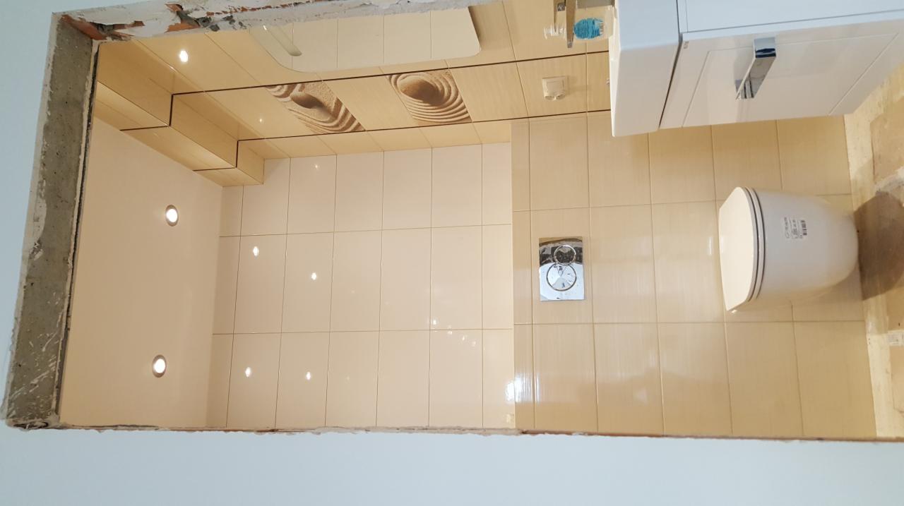 Wykończona łazienka z beżowymi płytkami, podwieszana toaleta, umywalka z szafką i oświetleniem punktowym w suficie podwieszanym.