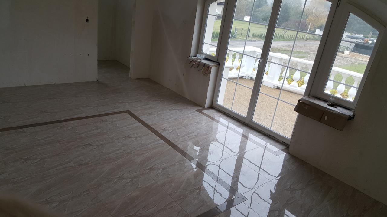 Ułożona podłoga z płytek ceramicznych w jasnych odcieniach z ciemniejszym obramowaniem, widoczne refleksy światła z okna balkonowego, surowe ściany w tle.