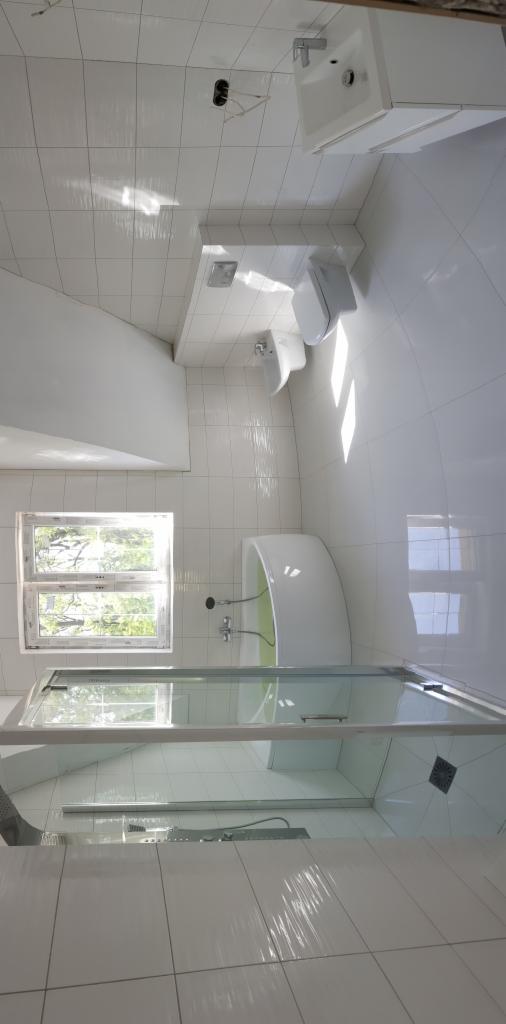 Wykończona łazienka w bieli: białe płytki na ścianach i podłodze, kabina prysznicowa ze szklanymi drzwiami, wanna, umywalka i toaleta. Widoczne okno z widokiem na zieleń.