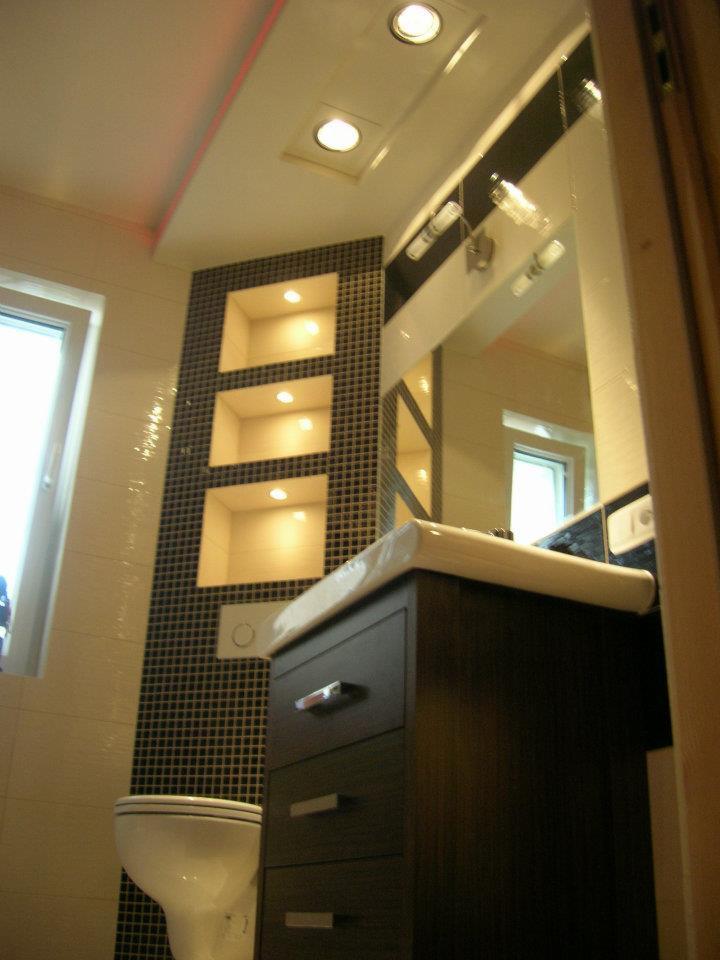 Wykończona łazienka z czarno-białą mozaiką, podświetlanymi niszami, umywalką z szafką w kolorze ciemnego drewna i wiszącą toaletą. Umywalka zabezpieczona folią ochronną.