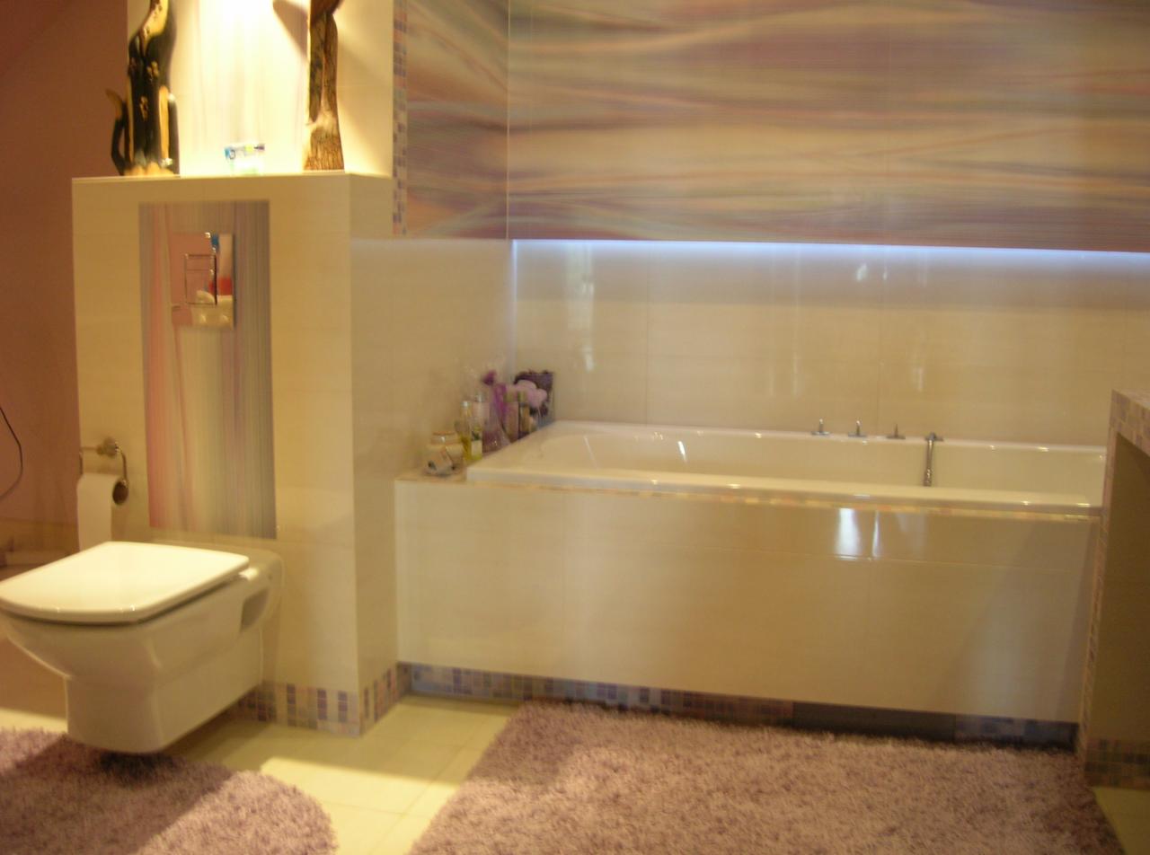 Jasna łazienka z wanną obudowaną płytkami, toaletą wiszącą i fioletowymi dywanikami, oświetlona listwą LED pod sufitem.