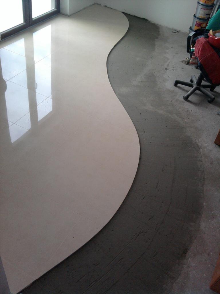 Układanie jasnych, beżowych płytek podłogowych w falistym wzorze, widoczny fragment podłoża przygotowanego pod płytki, w tle okno i puszki z farbą.