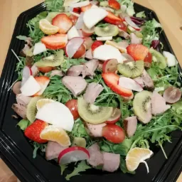 Gotowa sałatka z rukolą, kawałkami mięsa, kiwi, truskawkami, mandarynkami, rzodkiewką i winogronami, ułożona na czarnej, plastikowej tacy.