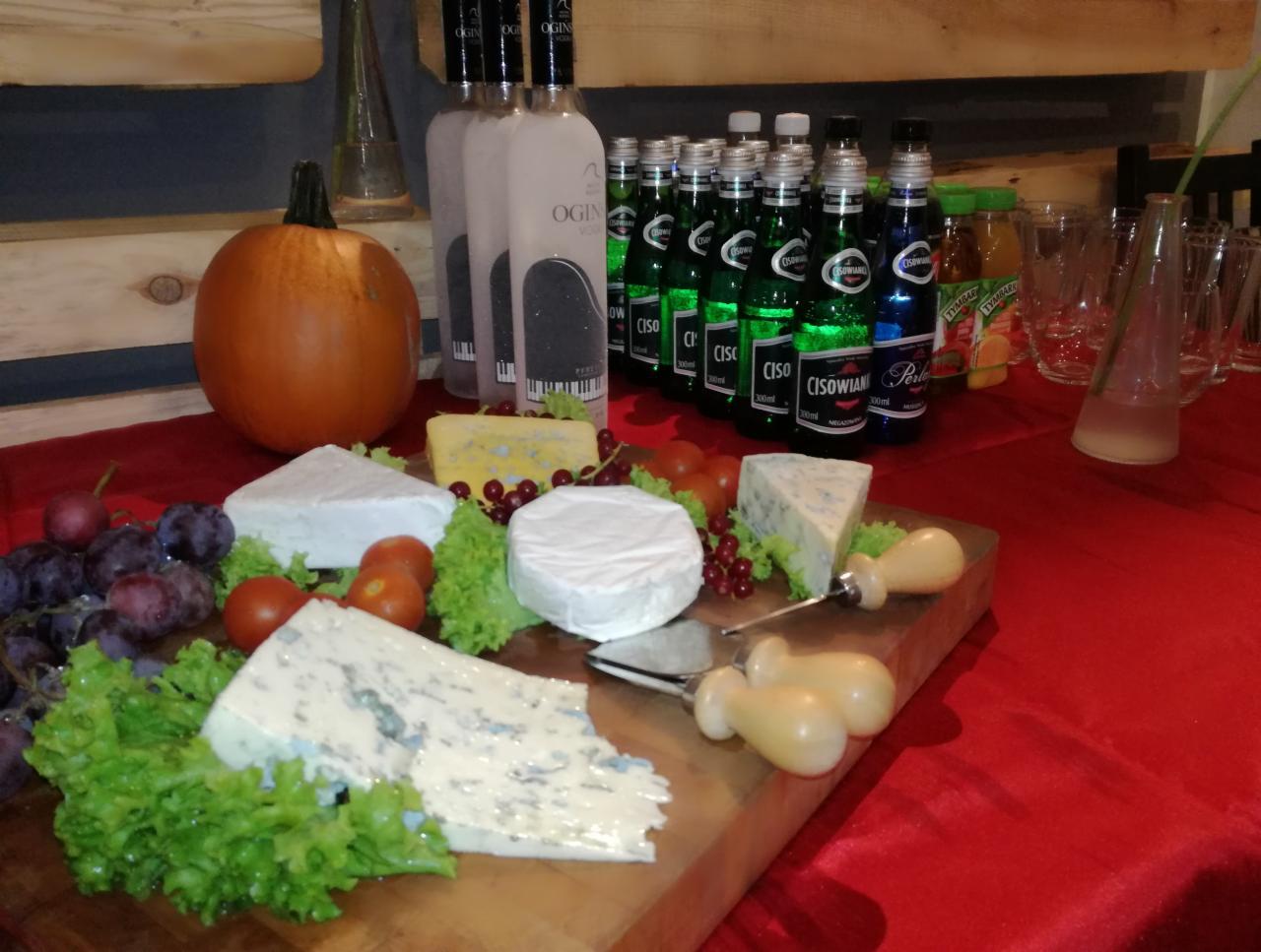 Stół z przekąskami: sery pleśniowe, winogrona, pomidorki koktajlowe, dynia, butelki wody Ciso i wódki Ogin, przygotowany na czerwonym obrusie.