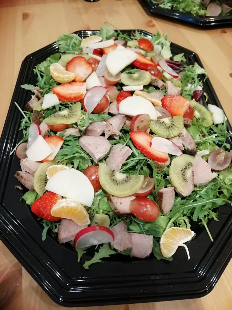 Gotowa sałatka z rukolą, kawałkami mięsa, kiwi, truskawkami, mandarynkami, rzodkiewką i winogronami, ułożona na czarnej, plastikowej tacy.