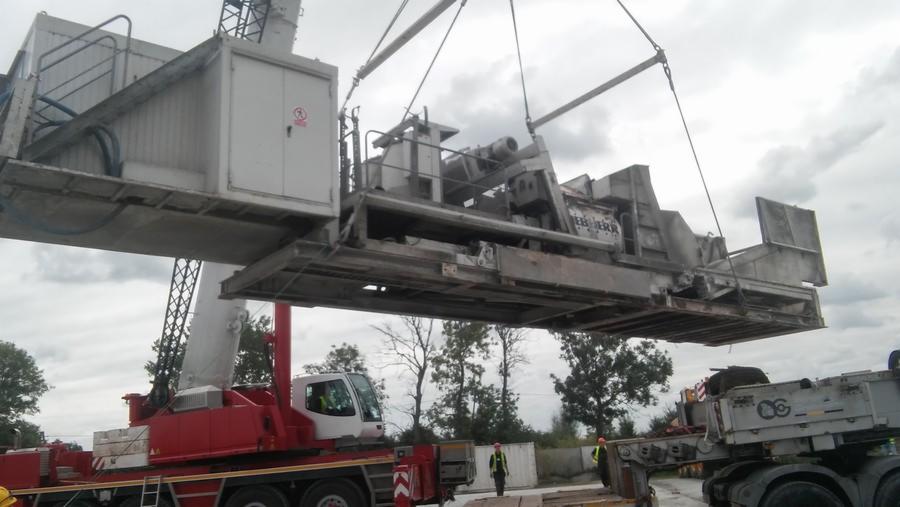 Ciężki, szary moduł maszyny budowlanej marki Liebherr podnoszony przez czerwony dźwig samochodowy na tle pochmurnego nieba i drzew.