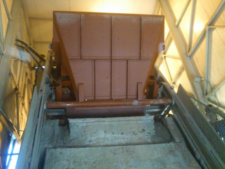 Widok z dołu na metalową konstrukcję przemysłową z elementami przypominającymi zsyp, wspartą na betonowej podstawie, w tle widoczne stalowe kratownice pod dachem hali.