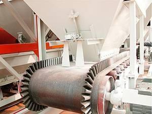 Fragment linii produkcyjnej z widocznym podajnikiem taśmowym z zębatymi rolkami, element konstrukcji pomalowany na biało i czerwono.