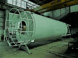 Duży, cylindryczny zbiornik przemysłowy z metalową konstrukcją wsporczą, umieszczony w hali produkcyjnej z widocznym suwnicą i oknami w tle.