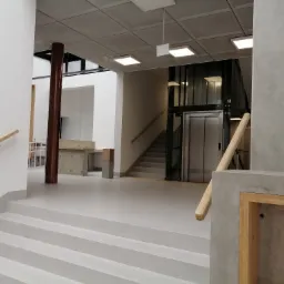 Jasnoszara posadzka jastrychowa w nowoczesnym budynku biurowym z widocznymi schodami, filarami i windą.