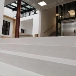 Nowoczesne wnętrze budynku z białymi schodami, przeszkloną windą i elementami konstrukcyjnymi z drewna i metalu, widok od dołu schodów.