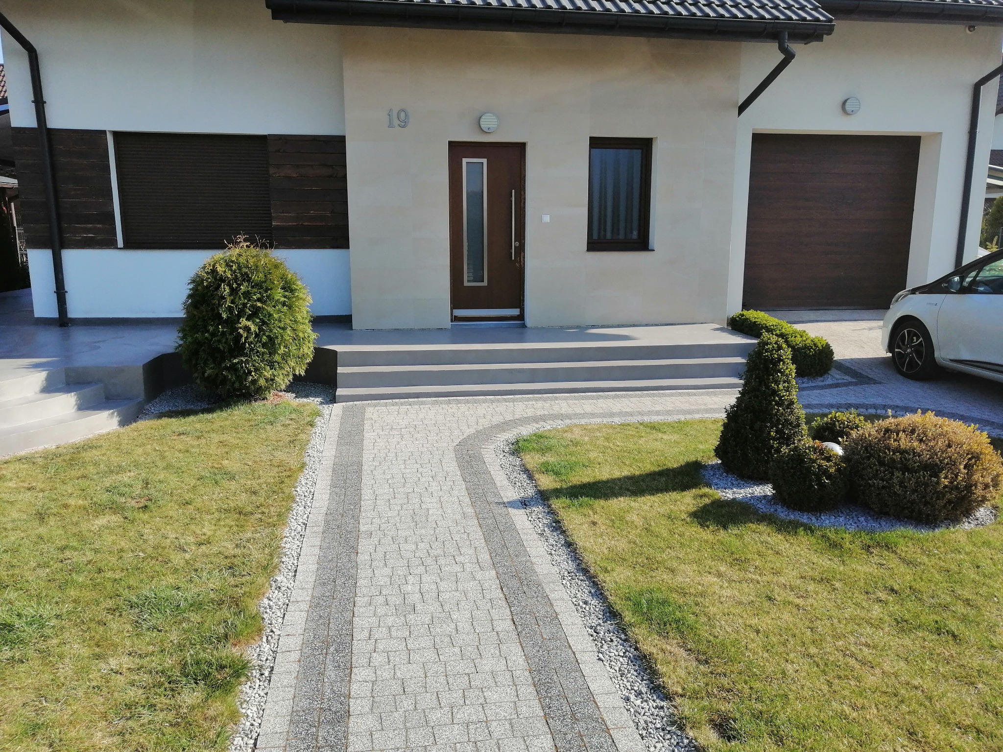 Nowoczesny dom jednorodzinny z jasną elewacją, ciemnobrązową stolarką okienną i drzwiową, oraz brukowanym chodnikiem z jasnoszarej kostki prowadzącym do wejścia, otoczony zielonym trawnikiem...