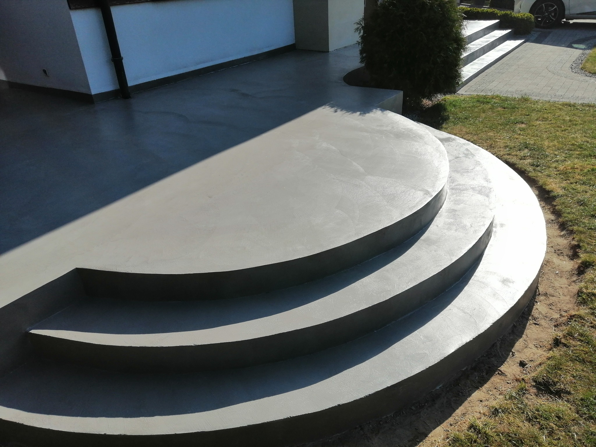 Półokrągłe, betonowe schody wejściowe z trzema stopniami prowadzące do domu, z widoczną szarą posadzką i fragmentem trawnika.